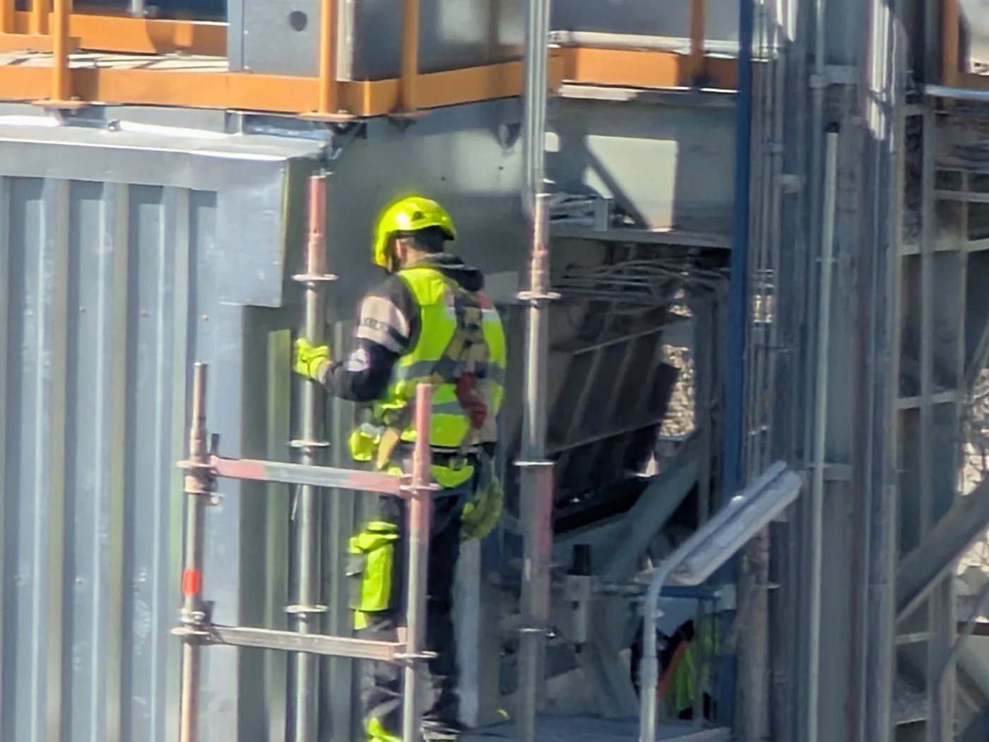 Pracovník v přilbě, reflexní vestě a postroji stoupá po lešení u průmyslového objektu