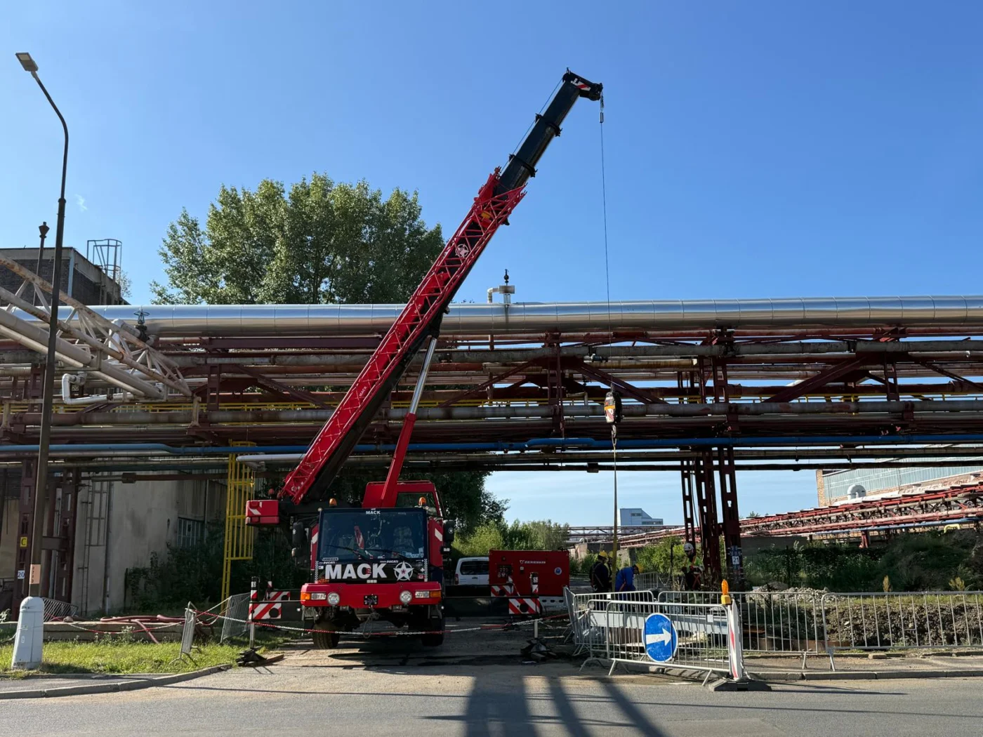 Autojeřáb pod potrubním mostem v průmyslovém areálu, pracoviště ohrazené zábradlím
